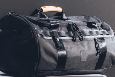 The Brown Buffalo Black duffel bag with orange handle on a gray background - Performance & Sustainability