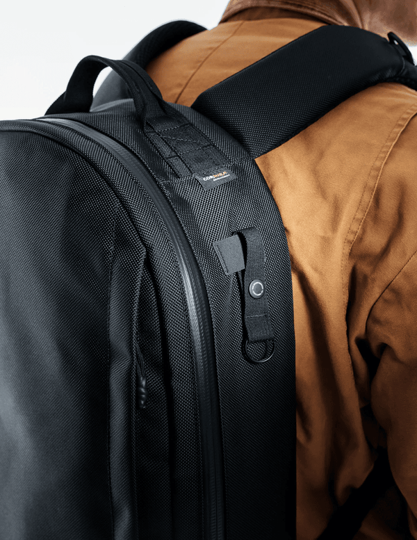The Brown Buffalo Black backpack worn by a person in a brown jacket on a white background